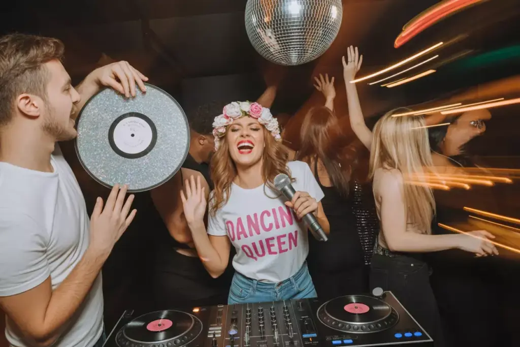 A woman in a Dancing Queen shirt sings at a lively party with a DJ and people dancing under a disco ball.