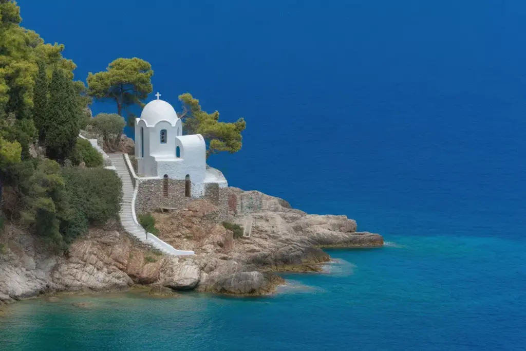 White chapel with a dome stands on rocky coastline, surrounded by trees, overlooking clear blue sea and sky.