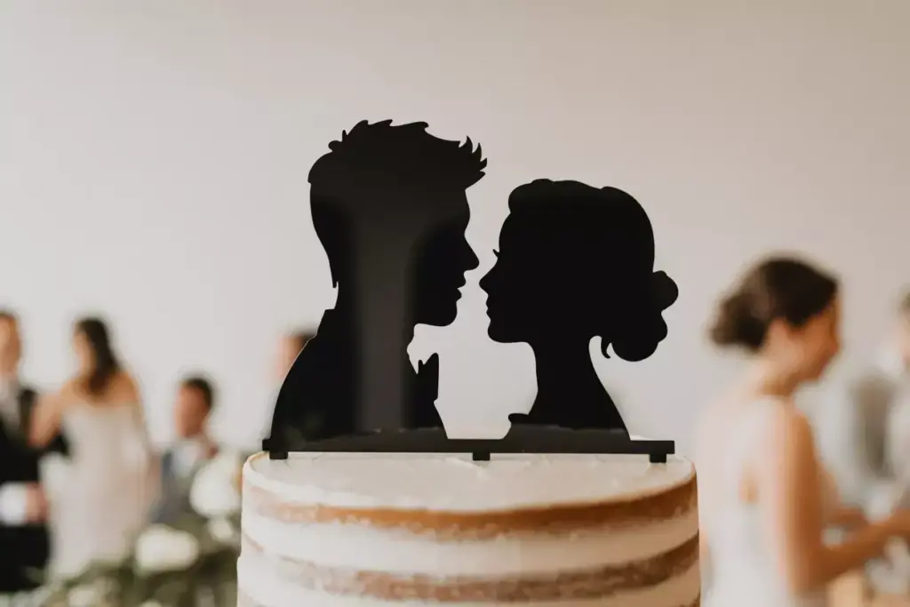 Silhouette cake topper of a couple facing each other on a tiered wedding cake, with guests blurred in background.