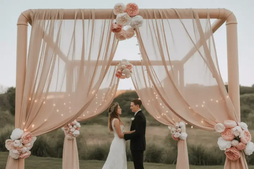 A couple stands under a pink floral wedding arch, holding hands and facing each other outdoors.