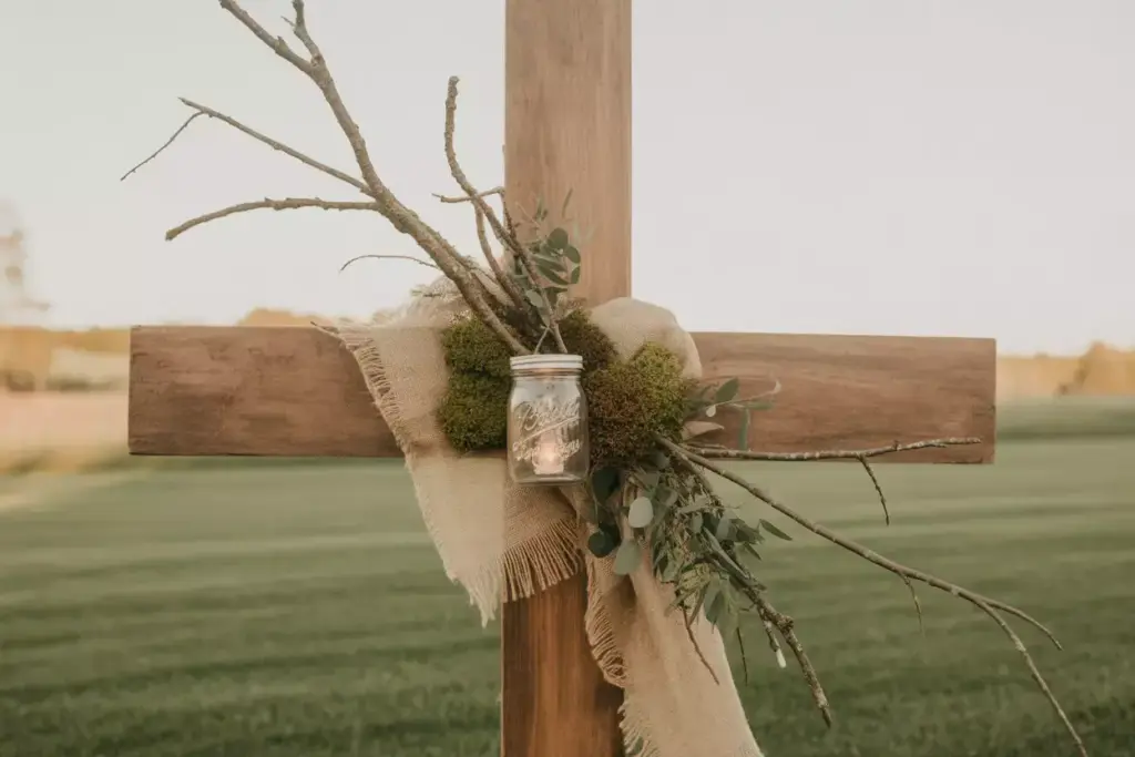 A wooden cross decorated with burlap, twigs, greenery, moss, and a mason jar in a grassy field.