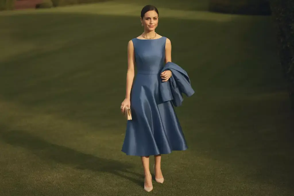 Woman in a sleeveless blue dress holding a clutch and jacket, standing on a grassy lawn.