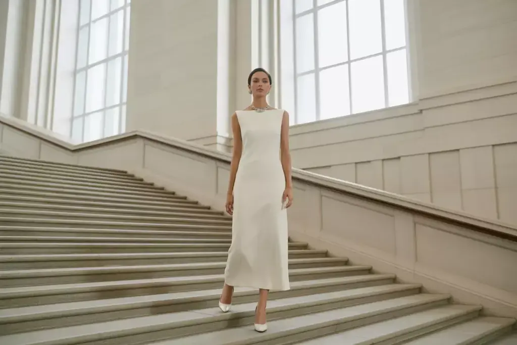 Woman in a sleeveless white dress and heels walking down elegant, wide stairs in a bright, grand hall.