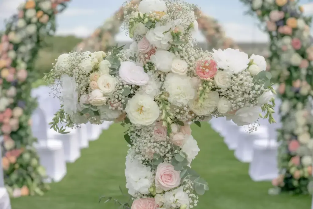 A cross made of white and pink flowers stands at an outdoor wedding ceremony with floral arches and chairs.