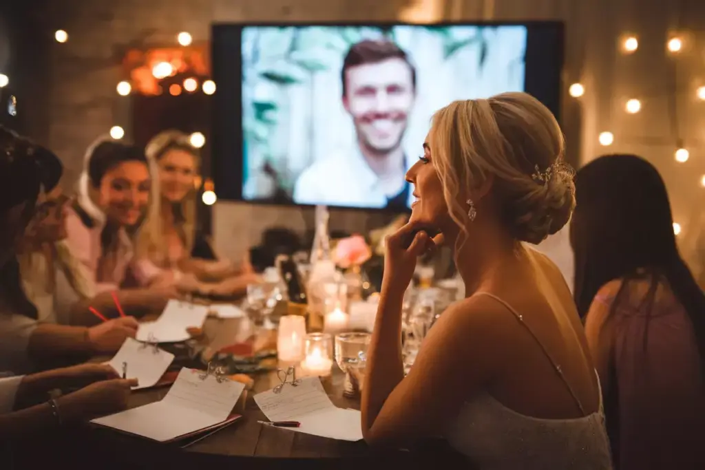 Bride smiles at a table during a wedding reception, with a man’s face on a screen in the background.