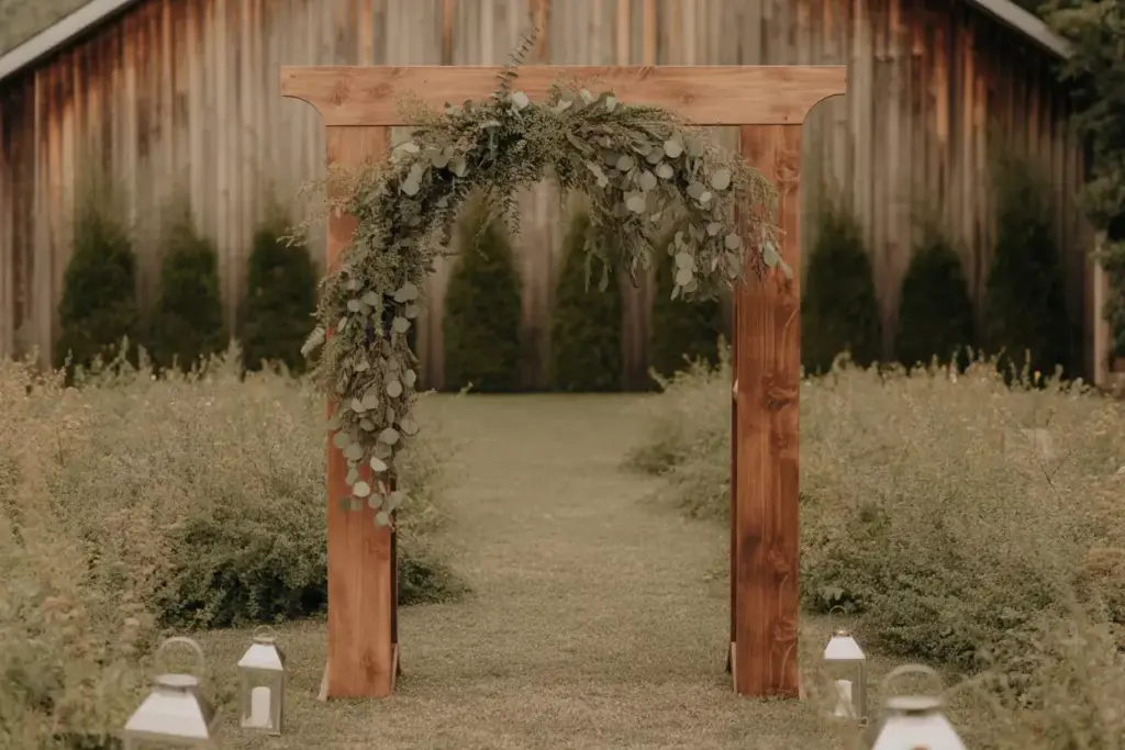 Rustic wooden wedding arch with greenery garland, surrounded by grass and lanterns, outdoors.