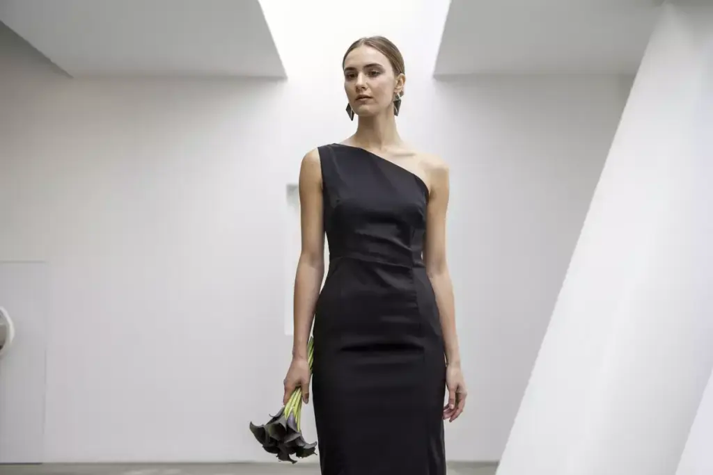 Woman in a sleek, one-shoulder black dress holding dark flowers, standing in a modern, white space.