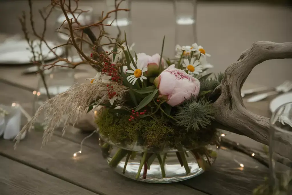 Glass bowl with pink peonies, daisies, moss, and twigs on a wooden table, next to driftwood and fairy lights.