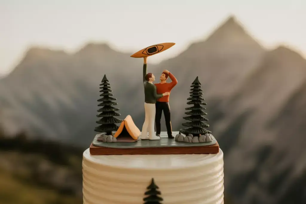 Cake topper with two figures holding a kayak, trees, and a tent, with mountains in the background.