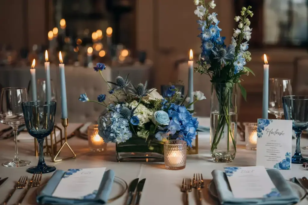 Elegant table setting with blue flowers, candles, glassware, and menus on a white tablecloth.