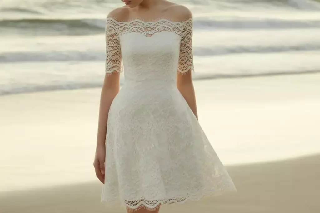 Woman in a short, off-the-shoulder lace wedding dress standing on a beach near the ocean.