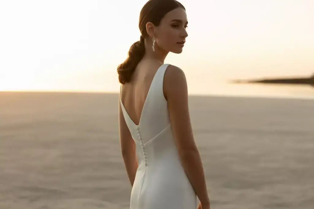 Woman in an elegant white dress stands on a beach at sunset, facing away from the camera.