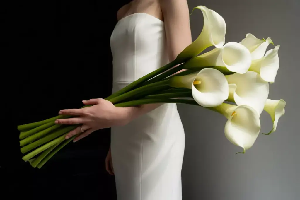 A person in a white dress holds a bouquet of white calla lilies against a dark background.