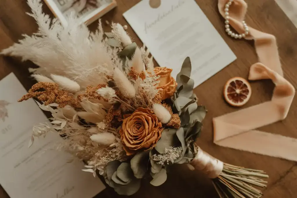 A rustic bouquet with dried flowers, wedding invitations, ribbon, pearls, and a dried orange slice on a table.