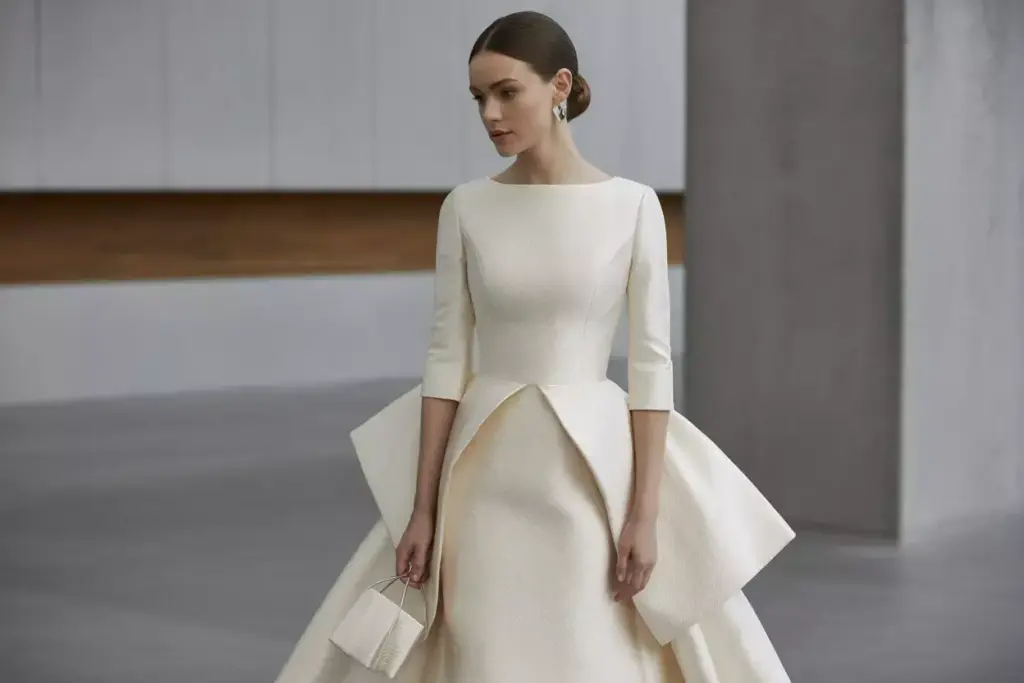 Woman in an elegant, modern white wedding dress with a sculptural skirt, holding a small matching purse.