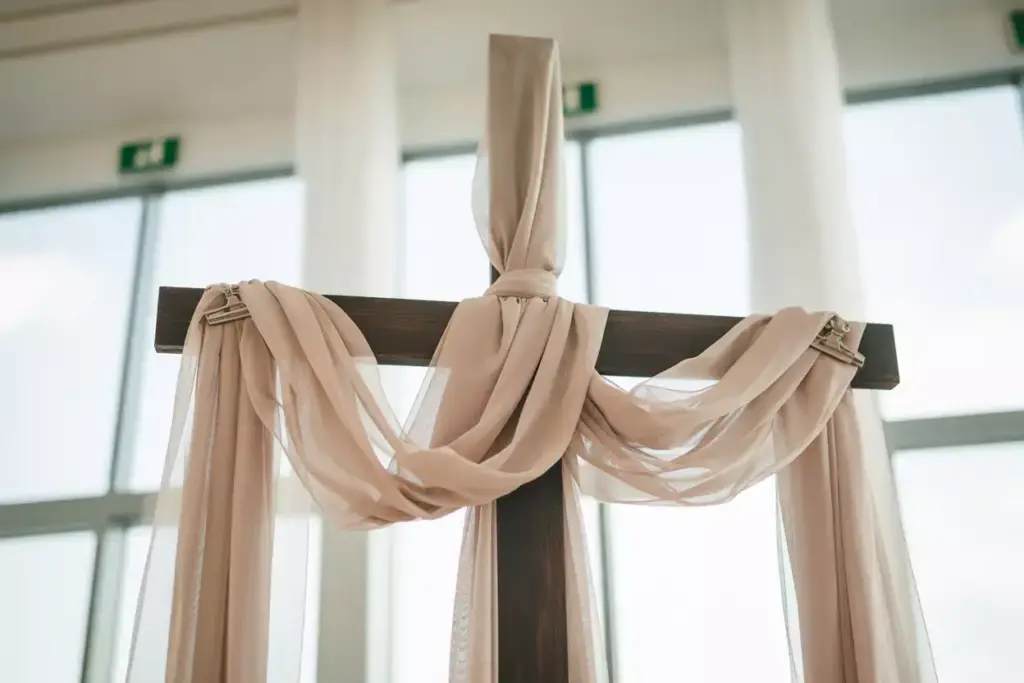 A wooden cross draped with light beige fabric, set in front of large windows with sheer curtains.