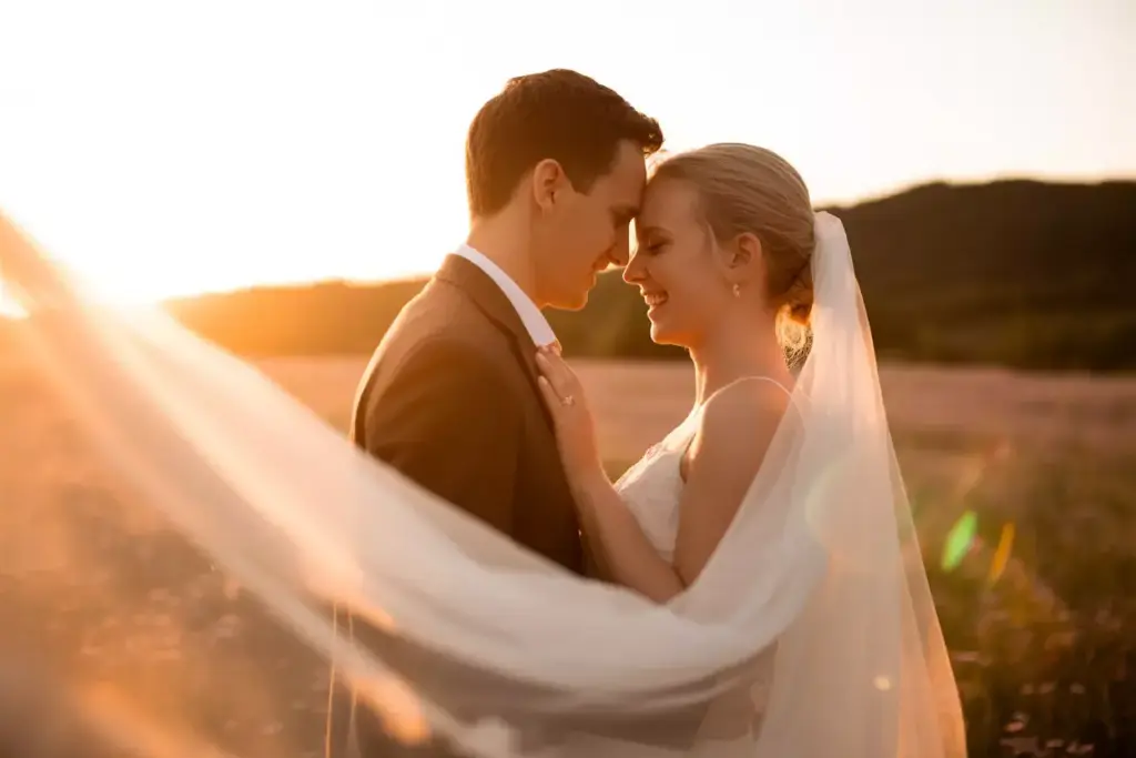 Bride and groom embrace in a sunlit field, smiling with their foreheads touching and veil flowing.