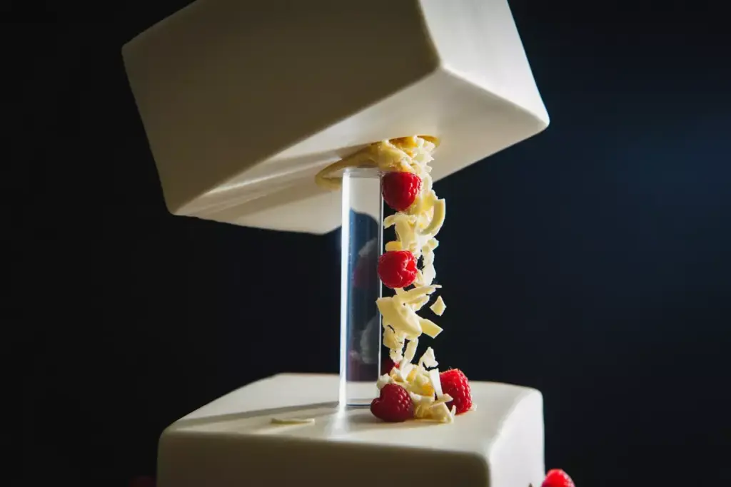 A modern square cake with raspberries and white chocolate cascading through a clear tube between two tiers.