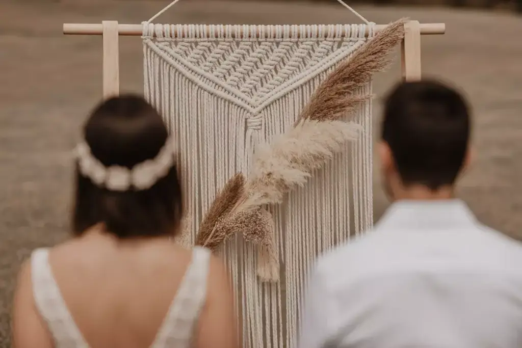 A couple faces a macramé wall hanging with pampas grass at an outdoor ceremony.