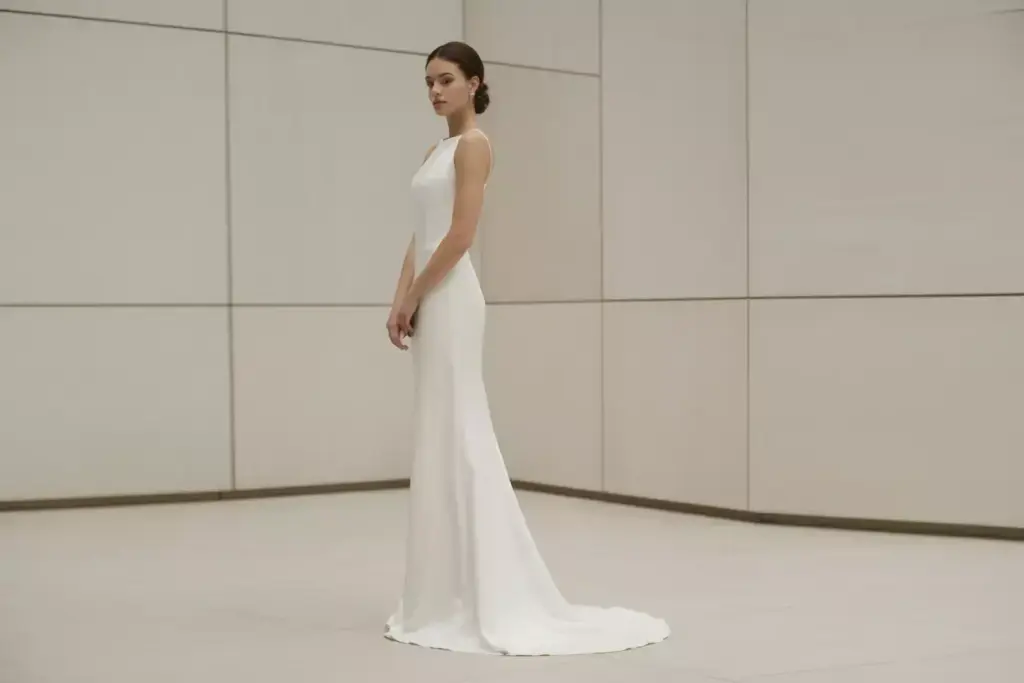Woman in a sleek, white sleeveless gown with a train, standing in a minimalist, light-colored room.