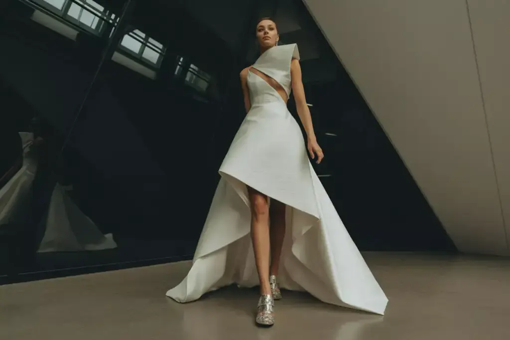 A woman in a modern, asymmetrical white gown and silver heels stands indoors against a dark geometric wall.