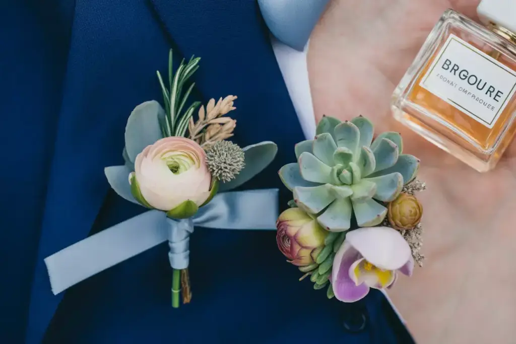 A hand holds a bottle of perfume next to floral boutonnieres pinned on a blue suit jacket.