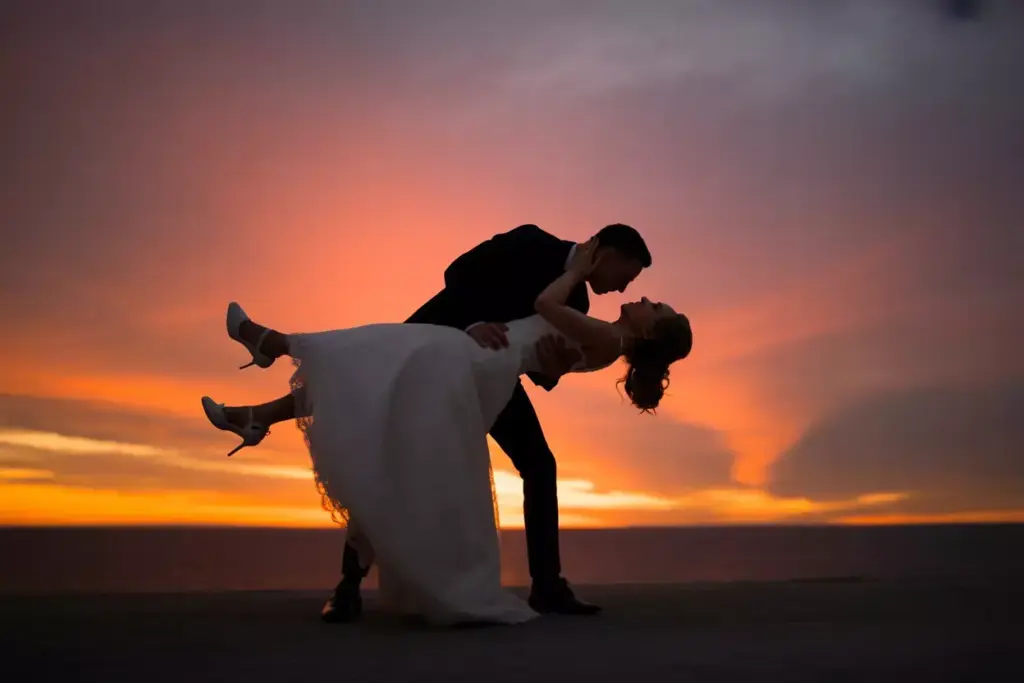 A couple dances at sunset, with the groom dipping the bride against a vibrant orange sky.