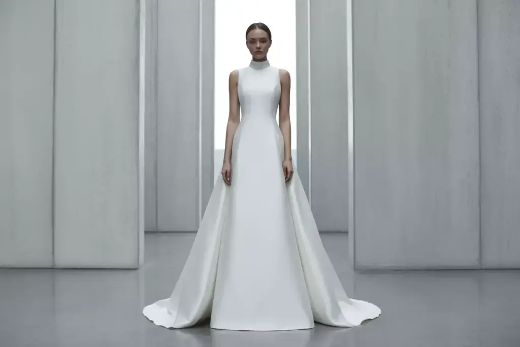 A woman in a sleeveless, high-neck white gown stands in a minimalist, modern room with tall grey walls.