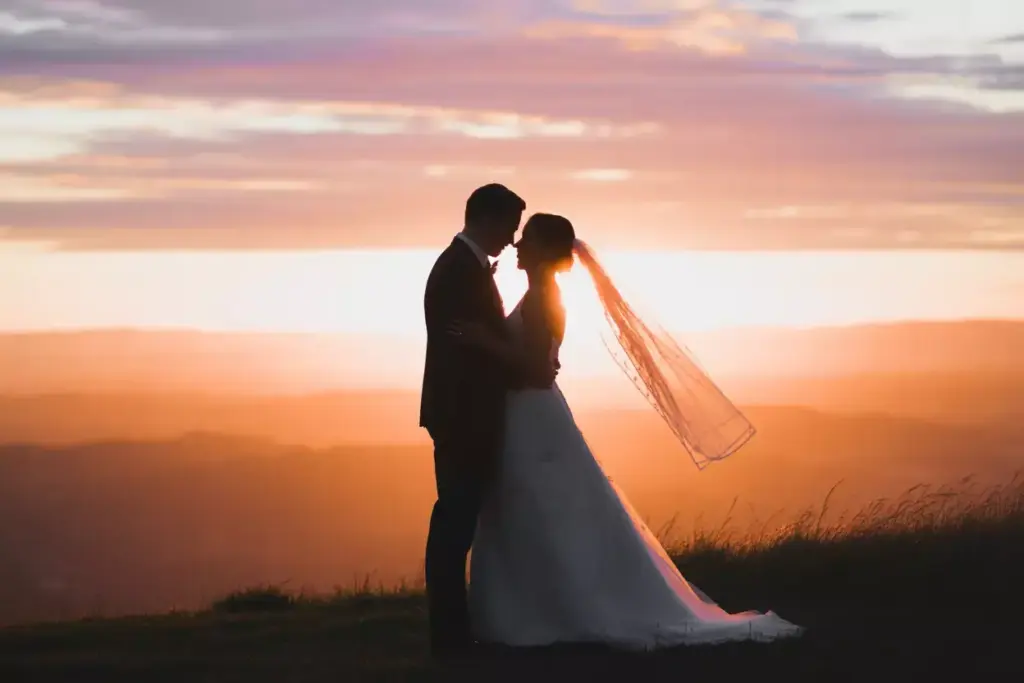 Bride and groom embrace at sunset on a hilltop, with a glowing sky and veil gently flowing.