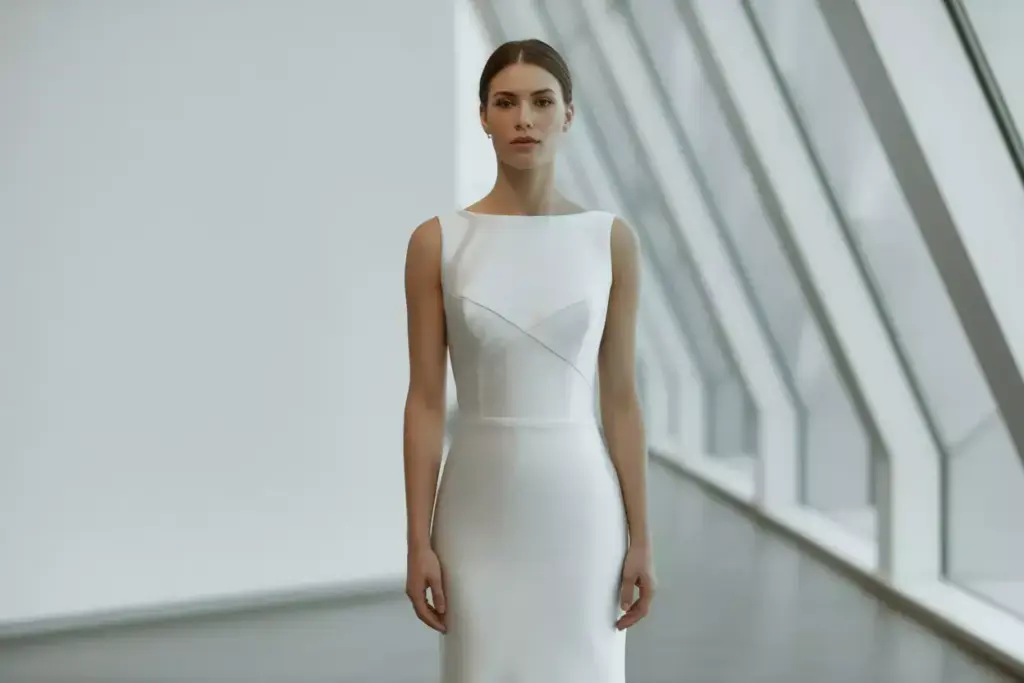 Woman in a sleeveless white dress stands in a modern, bright room with large angular windows.
