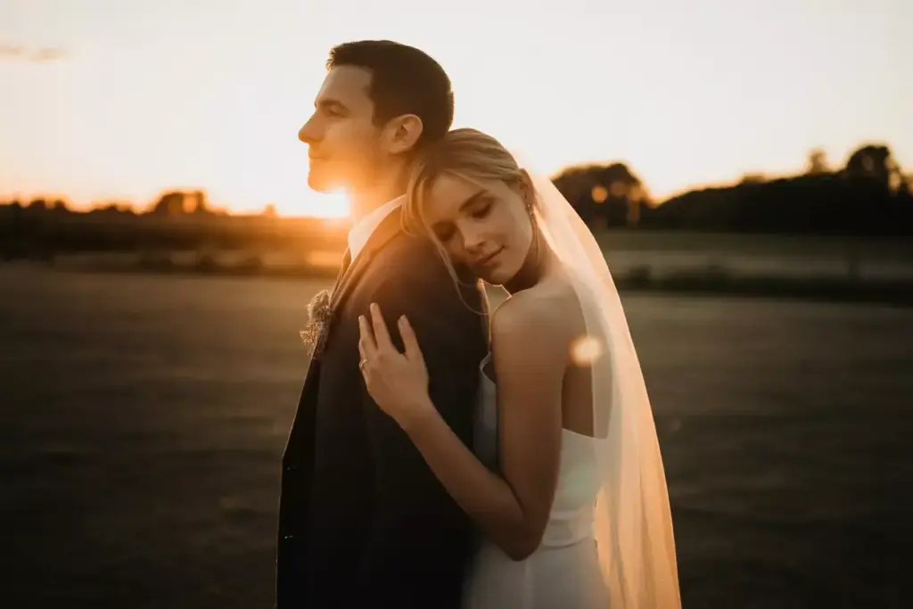 Bride hugs groom from behind at sunset in an open field, both wearing wedding attire and looking serene.