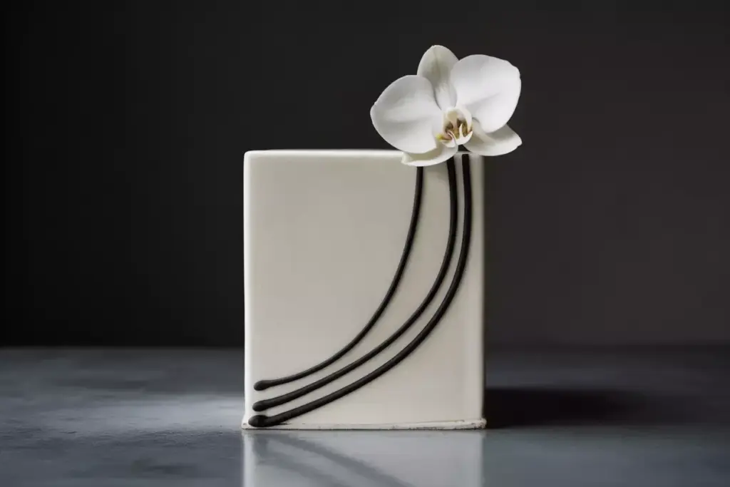 Square white cake with a white orchid and three curved black lines as decoration, on a gray surface.