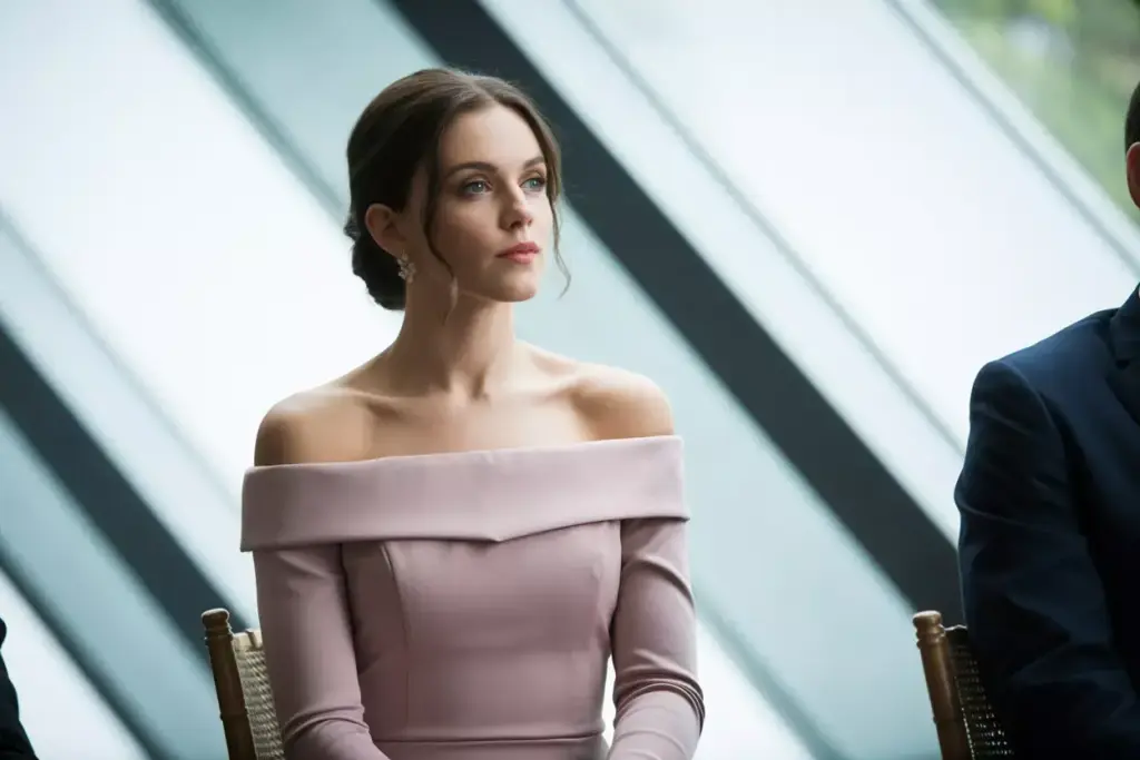 Woman in an off-shoulder light pink dress sits indoors, looking thoughtful, with modern windows behind her.