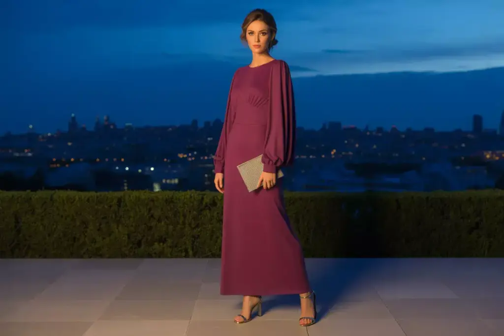 Woman in a long purple dress holding a clutch, standing outdoors at dusk with city lights in the background.