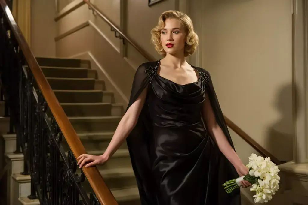 Woman in an elegant black gown holding white flowers stands by a staircase, looking thoughtfully to the side.