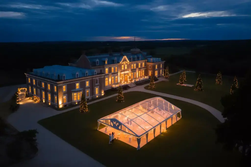 An elegant mansion with glowing lights and a transparent event tent on the lawn at dusk.
