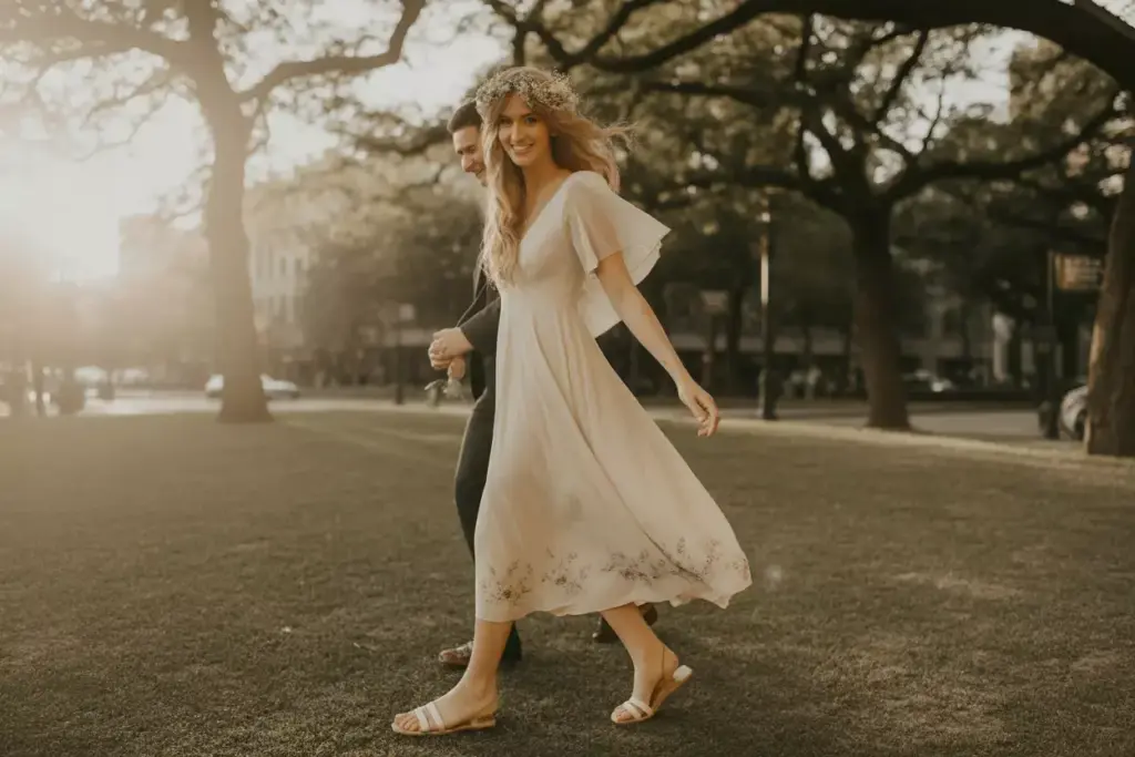 Woman in a white dress and sandals walks smiling in a sunlit park, holding hands with a man.