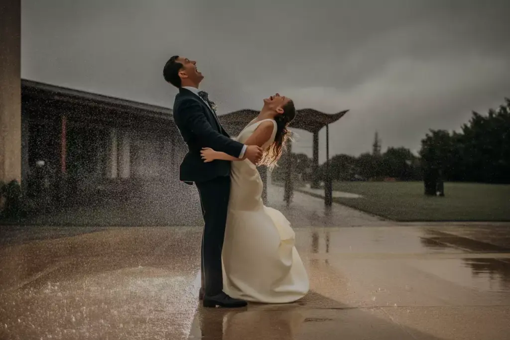 Bride and groom laugh joyfully in the rain on a wet patio, embracing each other in wedding attire.